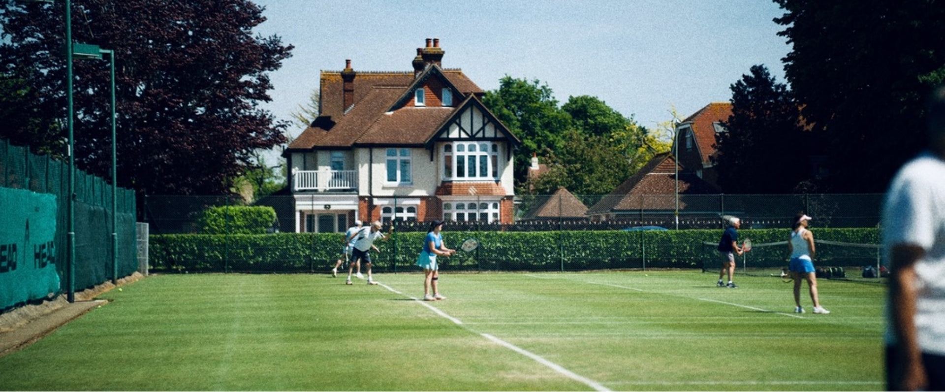 Menschen spielen auf Grasplätzen Tennis. Im Hintergrund ein Fachwerkhaus.