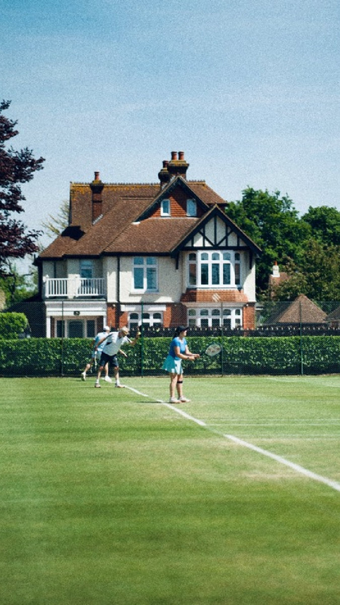 Menschen spielen auf Grasplätzen Tennis. Im Hintergrund ein Fachwerkhaus.