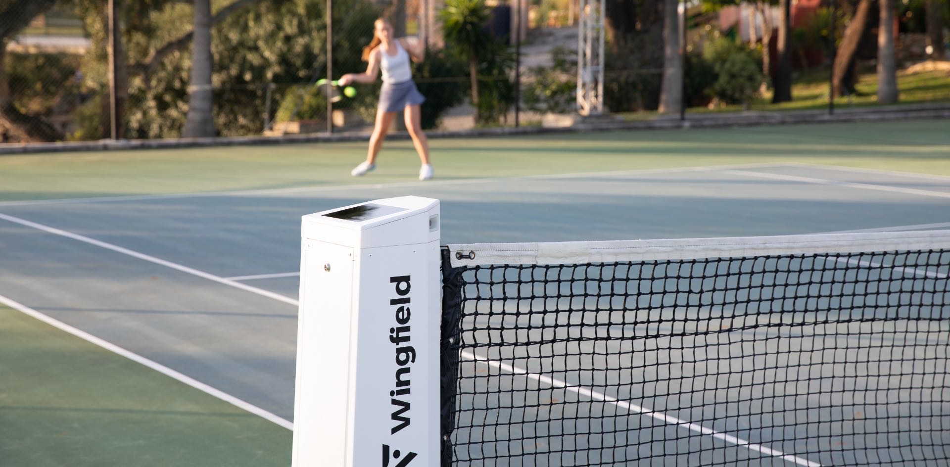 Die weiße Wingfield Box auf einem Hartplatz. Im Hintergrund eine Tennisspielerin.